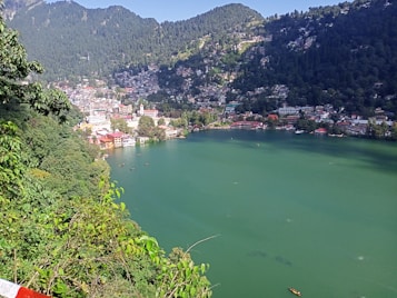 A serene lake surrounded by lush green hills with a densely populated town visible on the slopes. The clear sky suggests a bright and sunny day, while the vibrant greenery highlights the natural beauty of the area.