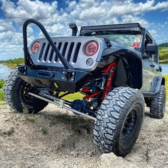 A rugged off-road vehicle with large tires and a lifted suspension is parked on uneven terrain. The vehicle is black with red suspension components visible. The sky is partly cloudy, and there is a body of water and vegetation in the background, indicating a natural outdoor setting.