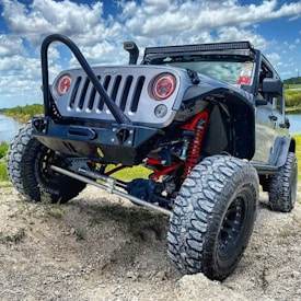 A rugged off-road vehicle with large tires and a lifted suspension is parked on uneven terrain. The vehicle is black with red suspension components visible. The sky is partly cloudy, and there is a body of water and vegetation in the background, indicating a natural outdoor setting.
