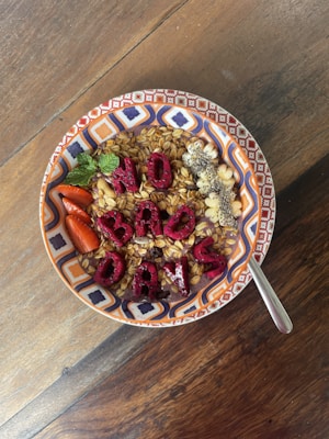A bowl of granola topped with various fruits and seeds, arranged neatly. The fruits include sliced strawberries and pieces of a fruit arranged to spell out the words 'NO BAD DAYS.' The bowl is patterned with colorful geometric designs, and a spoon is placed on the wooden table next to the bowl.