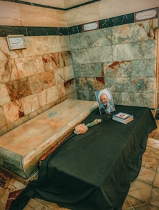 A marble tomb in a room with beautifully patterned stone walls. On top of the tomb, there is a black cloth draped overhanging the side. A framed photograph of a religious figure is placed atop the cloth along with a few books and a single bouquet of flowers.