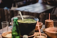 A rustic table setting featuring a large black pot filled with a green soup, garnished with herbs. The pot is accompanied by several clay bowls and cups, some containing drinks with straws, along with a green glass bottle and an empty glass. The atmosphere suggests a cozy and warm environment.