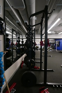 The image depicts a modern gym setting with various exercise equipment. In the foreground, there is a weightlifting rack with multiple weights stacked on both sides. The gym has mirrored walls reflecting the equipment and the spacious interior. Overhead, there are large industrial-style ventilation ducts and bright fluorescent lighting.
