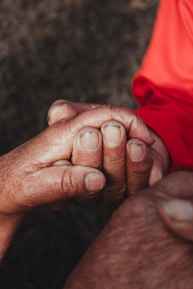 Two hands are gently clasped together, signifying a gesture of support or comfort. The image captures the textures and details of the skin, highlighting both age and tenderness. One hand wears an orange sleeve.