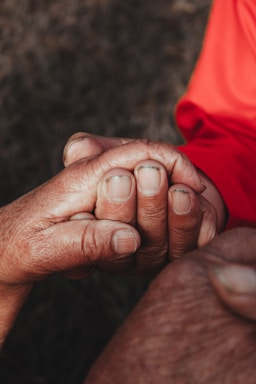 Two hands are gently clasped together, signifying a gesture of support or comfort. The image captures the textures and details of the skin, highlighting both age and tenderness. One hand wears an orange sleeve.