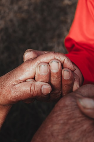 Two hands are gently clasped together, signifying a gesture of support or comfort. The image captures the textures and details of the skin, highlighting both age and tenderness. One hand wears an orange sleeve.