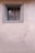 A small, barred window set in an unadorned stucco wall. The window frame is painted in a faded gray color, suggesting age and weathering. The wall has a light beige hue with a slightly textured surface.