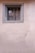 A small, barred window set in an unadorned stucco wall. The window frame is painted in a faded gray color, suggesting age and weathering. The wall has a light beige hue with a slightly textured surface.