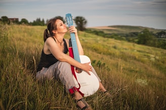 A woman sitting in a grassy field, leaning against a guitar. She appears relaxed, gazing into the distance. The landscape is expansive, with rolling hills under a calm sky.