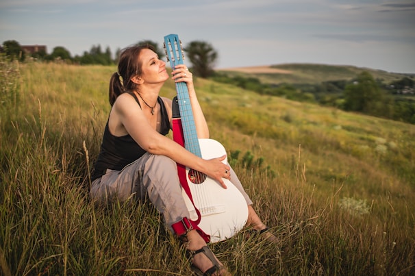 A woman sitting in a grassy field, leaning against a guitar. She appears relaxed, gazing into the distance. The landscape is expansive, with rolling hills under a calm sky.