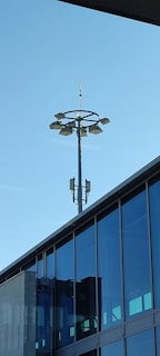 A modern building with large glass windows and a tall light pole structure extends into a clear blue sky. The light pole features multiple lamps arranged in a circular formation near the top and is equipped with additional equipment.