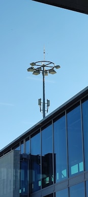 A modern building with large glass windows and a tall light pole structure extends into a clear blue sky. The light pole features multiple lamps arranged in a circular formation near the top and is equipped with additional equipment.