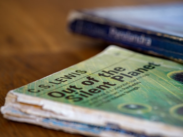 A close-up shot of a worn paperback book titled 'Out of the Silent Planet' by C.S. Lewis, resting on a wooden surface. In the background, the spine of another book is visible but out of focus.