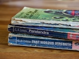 A stack of three worn paperback books is lying on a wooden surface. The books have visibly aged covers with scuffed edges and faded colors. They are authored by C.S. Lewis and include titles such as 'Out of the Silent Planet', 'Perelandra', and 'That Hideous Strength'. The top book has a green cover with abstract patterns, while the other two have predominantly blue covers.