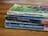 A stack of three worn paperback books is lying on a wooden surface. The books have visibly aged covers with scuffed edges and faded colors. They are authored by C.S. Lewis and include titles such as 'Out of the Silent Planet', 'Perelandra', and 'That Hideous Strength'. The top book has a green cover with abstract patterns, while the other two have predominantly blue covers.
