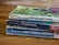 A stack of three worn paperback books is lying on a wooden surface. The books have visibly aged covers with scuffed edges and faded colors. They are authored by C.S. Lewis and include titles such as 'Out of the Silent Planet', 'Perelandra', and 'That Hideous Strength'. The top book has a green cover with abstract patterns, while the other two have predominantly blue covers.
