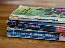 A stack of three worn paperback books is lying on a wooden surface. The books have visibly aged covers with scuffed edges and faded colors. They are authored by C.S. Lewis and include titles such as 'Out of the Silent Planet', 'Perelandra', and 'That Hideous Strength'. The top book has a green cover with abstract patterns, while the other two have predominantly blue covers.