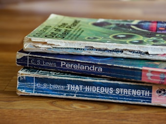A stack of three worn paperback books is lying on a wooden surface. The books have visibly aged covers with scuffed edges and faded colors. They are authored by C.S. Lewis and include titles such as 'Out of the Silent Planet', 'Perelandra', and 'That Hideous Strength'. The top book has a green cover with abstract patterns, while the other two have predominantly blue covers.