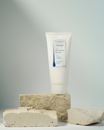 A tube of soft make-up remover labeled Oleogel is prominently displayed, standing upright on a stack of rough, light-colored stone blocks, against a softly lit neutral background.