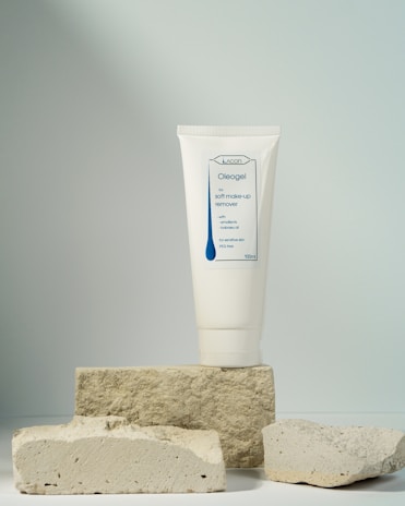 A tube of soft make-up remover labeled Oleogel is prominently displayed, standing upright on a stack of rough, light-colored stone blocks, against a softly lit neutral background.