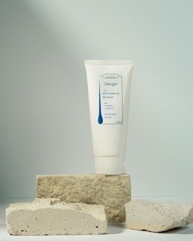 A tube of soft make-up remover labeled Oleogel is prominently displayed, standing upright on a stack of rough, light-colored stone blocks, against a softly lit neutral background.