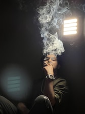 A person is sitting in a dimly lit room with a contemplative expression, exhaling a large plume of smoke. The light from a rectangular source creates a dramatic effect, with shadows and smoke adding to the atmosphere.