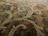 An intricately patterned carpet featuring swirling, abstract designs with an ornate floral-style motif. The fibers show a blend of colors including earthy tones, creating a textured and rich appearance.