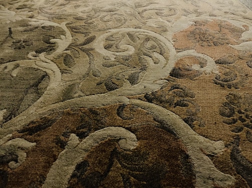An intricately patterned carpet featuring swirling, abstract designs with an ornate floral-style motif. The fibers show a blend of colors including earthy tones, creating a textured and rich appearance.