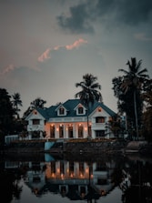 A large, elegant house with multiple gables and well-lit windows is surrounded by palm trees, situated beside a calm body of water that reflects the house and trees. The sky is overcast with dark clouds, adding a dramatic atmosphere.