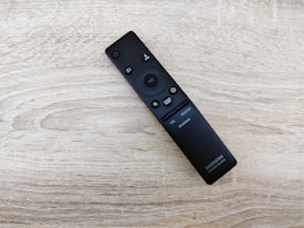 A sleek black remote control is placed on a light wooden surface. The remote has several buttons, including a power button, directional buttons, and sound mode options. The surface has a textured, natural wood grain pattern.