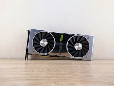 A metallic computer graphics card rests on a wooden surface. It features two large black cooling fans and a label indicating 'RTX 2080 Super'. The card has a sleek, modern design with silver and black elements.