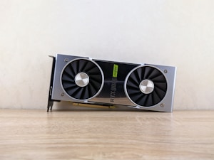 A metallic computer graphics card rests on a wooden surface. It features two large black cooling fans and a label indicating 'RTX 2080 Super'. The card has a sleek, modern design with silver and black elements.
