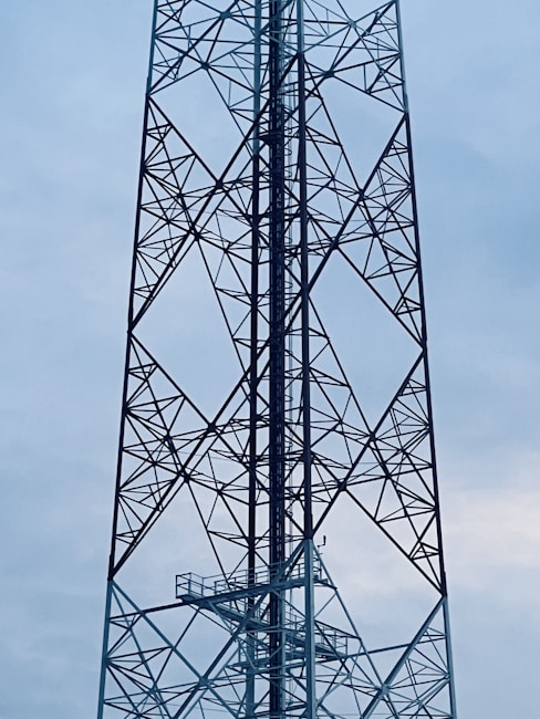 A tall metal transmission tower stands against a cloudy sky. The structure features a lattice framework, composed of interconnected steel beams, creating a geometric pattern of triangles and squares.