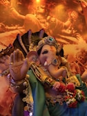 A detailed and ornate sculpture of a deity with an elephant head, adorned with jewels and colorful garments. The deity's pose includes an upright palm with symbols and an expression of calm and peace. Background features warm, glowing imagery of another spiritual figure.