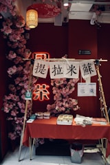 A small market stall or booth with a red tablecloth draped over the table, set against a vibrant red wall adorned with pink flower decorations. There are various items on the table, including food containers and utensils. Above the table, there are white banners with black text hanging on a wooden frame. A paper lantern and other hanging decorations add to the aesthetic.