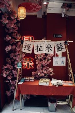 A small market stall or booth with a red tablecloth draped over the table, set against a vibrant red wall adorned with pink flower decorations. There are various items on the table, including food containers and utensils. Above the table, there are white banners with black text hanging on a wooden frame. A paper lantern and other hanging decorations add to the aesthetic.