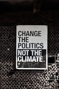 A large sign with bold, black and white text that reads 'Change the Politics Not the Climate' is affixed to a lattice-like, textured wall. The background has a dark, industrial feel.