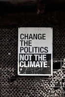 A large sign with bold, black and white text that reads 'Change the Politics Not the Climate' is affixed to a lattice-like, textured wall. The background has a dark, industrial feel.