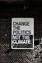A large sign with bold, black and white text that reads 'Change the Politics Not the Climate' is affixed to a lattice-like, textured wall. The background has a dark, industrial feel.