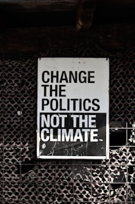 A large sign with bold, black and white text that reads 'Change the Politics Not the Climate' is affixed to a lattice-like, textured wall. The background has a dark, industrial feel.