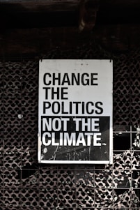 A large sign with bold, black and white text that reads 'Change the Politics Not the Climate' is affixed to a lattice-like, textured wall. The background has a dark, industrial feel.
