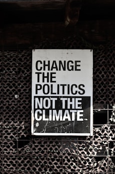 A large sign with bold, black and white text that reads 'Change the Politics Not the Climate' is affixed to a lattice-like, textured wall. The background has a dark, industrial feel.