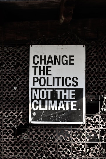 A large sign with bold, black and white text that reads 'Change the Politics Not the Climate' is affixed to a lattice-like, textured wall. The background has a dark, industrial feel.