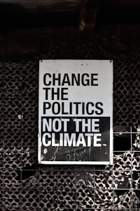 A large sign with bold, black and white text that reads 'Change the Politics Not the Climate' is affixed to a lattice-like, textured wall. The background has a dark, industrial feel.