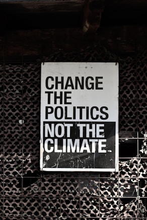 A large sign with bold, black and white text that reads 'Change the Politics Not the Climate' is affixed to a lattice-like, textured wall. The background has a dark, industrial feel.