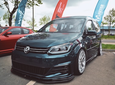 A modified Volkswagen van with sleek, low-profile tires is parked on a street. It is accompanied by a red car parked nearby. Flags bearing logos and text are placed behind them. The scene is set outdoors with some trees and a cloudy sky.