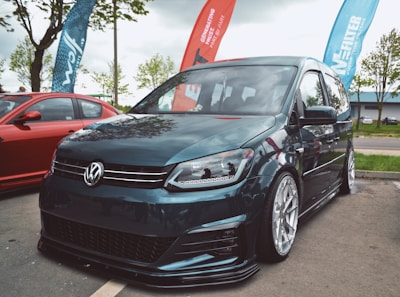 A modified Volkswagen van with sleek, low-profile tires is parked on a street. It is accompanied by a red car parked nearby. Flags bearing logos and text are placed behind them. The scene is set outdoors with some trees and a cloudy sky.