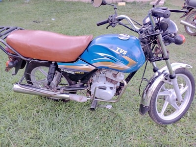 A motorbike with a blue and gray fuel tank, a brown seat, and black handlebars resting on green grass. The word 'TVS' is visible on the fuel tank.