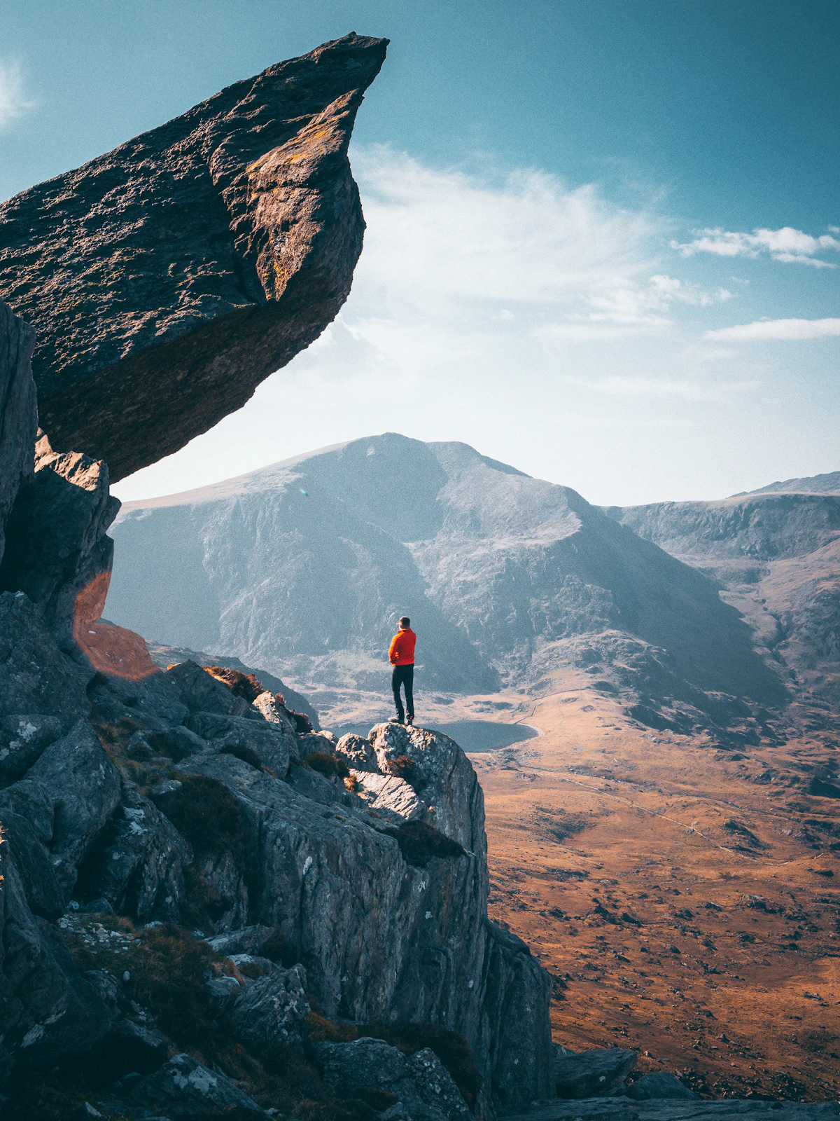 A hiker on a rock outcrop near Tryfan, under an overhanging slab, with a bare mountain and a small lake beyond