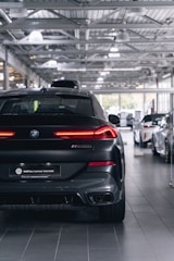 A sleek, black BMW M50i is parked in a modern, well-lit car showroom. The car's rear end faces the viewer, highlighting its distinctive taillights and sporty design. Other cars are visible in the background, and the ceiling features industrial-style lighting and structure.